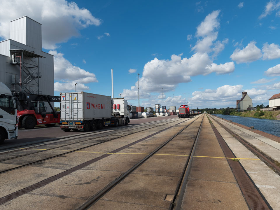 Blick auf einen Umschlagplatz mit mehreren Lkw, Eisenbahnschienen und einem Wasserkanal unter blauem Himmel mit Wolken