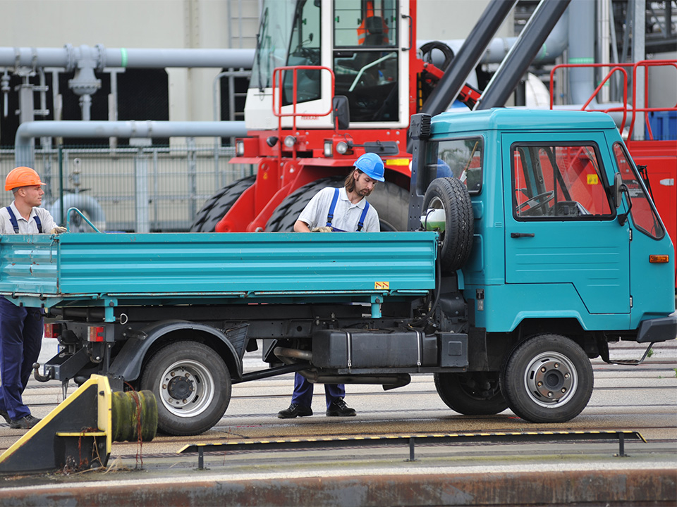 Zwei Männer mit Schutzhelmen stehen neben einem kleinen blauen Lastwagen auf einem Industriegelände