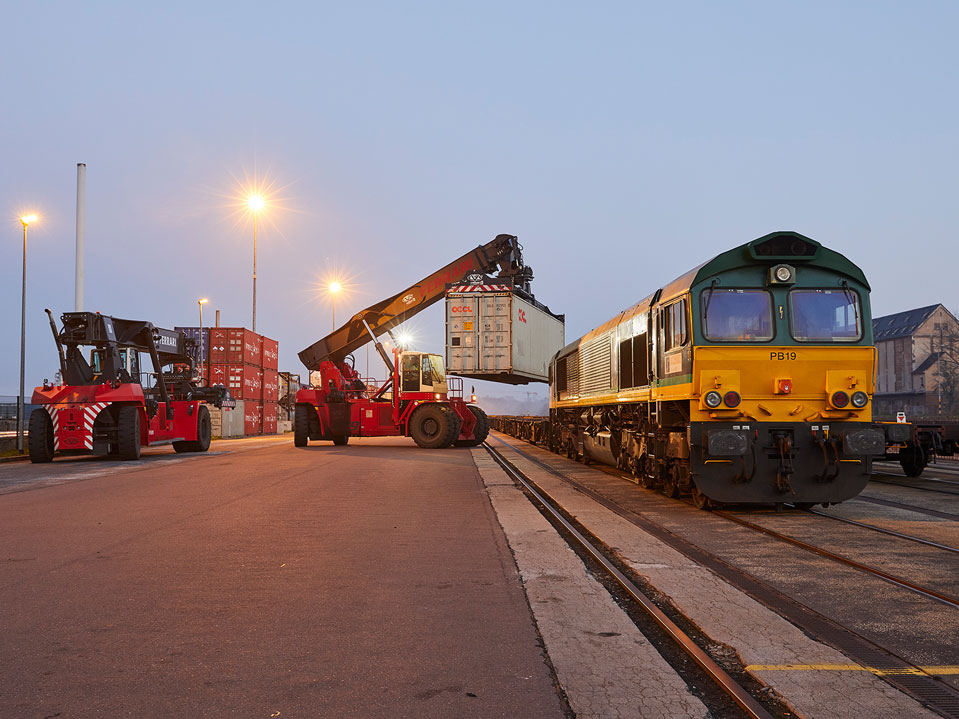 Zwei Containerstapler verladen einen Container neben einer wartenden Lokomotive auf einem Güterbahnhof bei Dämmerung