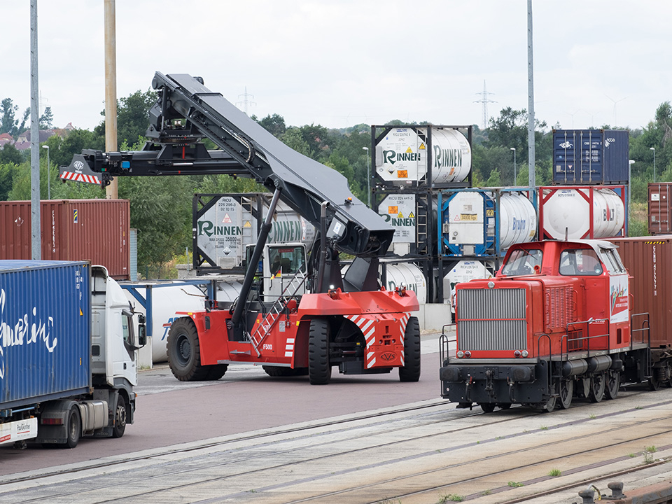 roter Containerstapler neben Lkw auf einem Umschlagplatz mit mehreren Containern und einer roten Rangierlokomotive auf Gleisen
