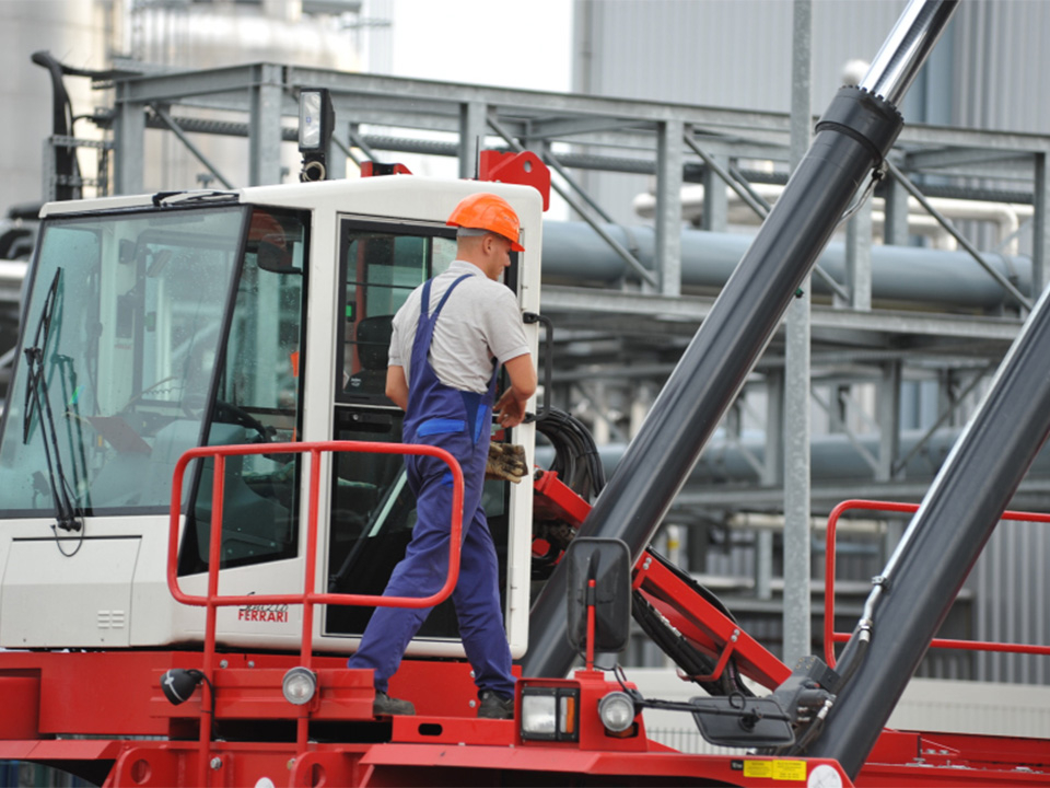Arbeiter mit orangem Schutzhelm und blauer Arbeitskleidung steht auf rotem Containerterminalfahrzeug
